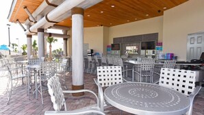 Ein Außenbereich mit einer Terrasse, Metallstühlen und -tischen, einer Bar mit Fernseher und Barkitchen, sowie einer Holzbalkendecke.