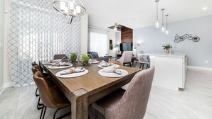 Uma sala de jantar moderna com uma mesa de madeira posta para quatro pessoas, cercada por cadeiras. Uma cozinha é visível ao fundo, com bancada branca e pia.