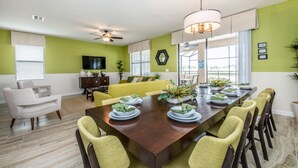 A dining area with a long table set for six, surrounded by yellow chairs. There is a ceiling fan, a television on the wall, and a window with blinds. - Your Beautiful Mansion Close to Disney on the Prestigious Champions Gate Resort, Orlando Mansion 2508 (Davenport)