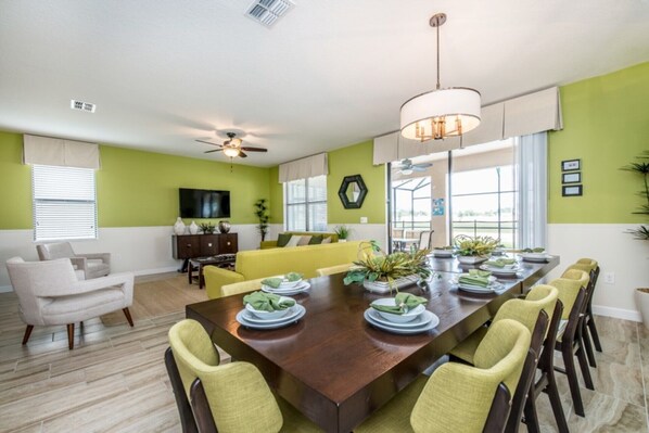 Une salle à manger avec une longue table dressée pour six personnes, entourée de chaises jaunes. On y trouve un ventilateur de plafond, un téléviseur accroché au mur et une fenêtre avec des stores.