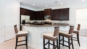 A modern kitchen with dark wood cabinets, a stainless steel refrigerator, a microwave, and a granite countertop. There are four bar stools with woven seats. - 5 Star Villa With Private Pool on Reunion Resort and Spa, Orlando Villa 4591 (Kissimmee)