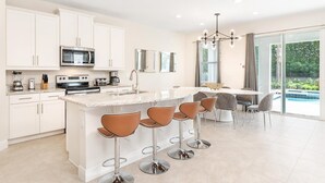 Una cocina moderna con armarios blancos, una isla central con taburetes y un microondas integrado, además de un comedor con vistas a una piscina.