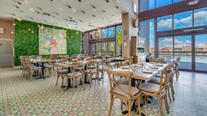 A spacious dining area with multiple tables and chairs, a green wall with artwork, and large windows offering a view of the outside.