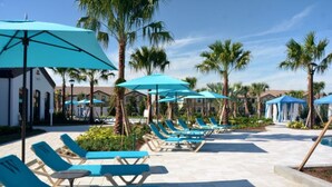 A poolside area with blue lounge chairs and umbrellas, surrounded by palm trees and buildings. - Beautiful Townhome Located on Windsor at Westside Resort, Orlando Townhome 5527 (Kissimmee)