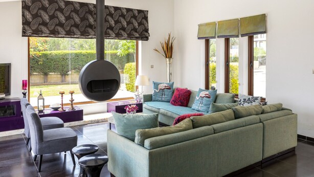 A modern living room with a large sofa, a coffee table, and a fireplace. There are large windows with blinds and a view of greenery outside. - At Last You can Rent the Perfect Luxury Villa in Protaras Close to all the Action, Protaras Villa 1569 (Paralimni)