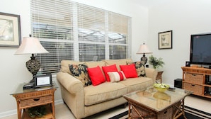 A living room with a beige sofa, red and black patterned cushions, two side tables with lamps, a glass coffee table, a television on a wooden stand, and framed artwork on the walls. - Beautiful 5 Star Villa With Private Pool on the Prestigious Windsor Hills Resort, Orlando Villa 4766 (Kissimmee)