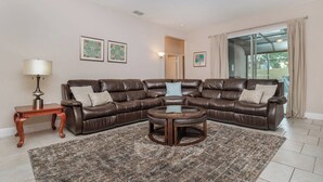 Un salon avec un canapé d’angle en cuir marron, une petite table d’appoint en bois, une table basse ronde et un tapis à motifs. Deux tableaux encadrés ornent le mur, et une fenêtre est agrémentée de rideaux.