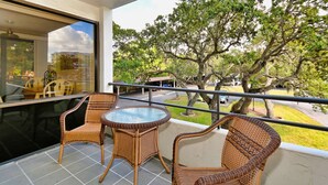 A balcony with wicker furniture, a glass table, and a view of trees and a road. - Beautiful Apartment on The Anchorage, Siesta Key Apartment 1006 (Siesta Key)