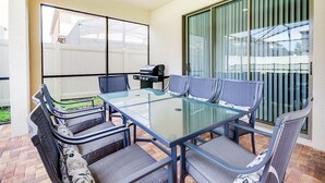 A patio with a glass-top table and chairs, a grill, and a view of a white fence and greenery. - Modern Oasis With 7 Bedrooms & Garden 5524 (Kissimmee)