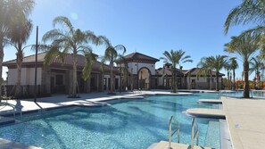 Une grande piscine extérieure dotée de plusieurs couloirs, entourée de palmiers et bordée d’un bâtiment au toit recouvert de tuiles.