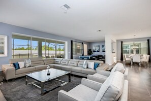 A modern living room with a sectional sofa, a glass coffee table, and a dining area with a table and chairs. - Beautiful Mansion With Private Pool, Close to Disney, Orlando Mansion 3128 (Kissimmee)