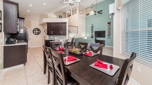 A modern kitchen with dark cabinetry, a dining area with a wooden table set for four, and a living area with a TV and artwork on the wall. - Exclusive Villa With Large Private Pool on Windsor Palms Resort, Orlando Villa 4884 (Kissimmee)