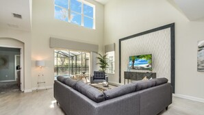 A spacious living room with a large gray sectional sofa, a flat-screen TV mounted on a textured wall, and a large window providing natural light. - Luxury 5 Star Villa on Storey Lake Resort,minutes From Disney World, Orlando Villa 3654 (Kissimmee)