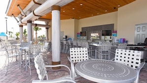 Un espace extérieur aménagé en terrasse, avec des chaises et des tables en métal, un coin bar équipé d’un téléviseur et de tout le matériel nécessaire, et une structure de plafond en bois.