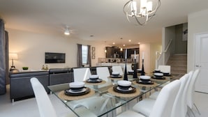 Uma sala de jantar moderna com mesa de vidro, cadeiras brancas e um lustre central.
