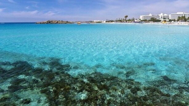 A coastal beach scene with clear turquoise water, rocky underwater terrain, and a distant shoreline with buildings.