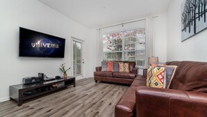 Een moderne woonkamer met een flatscreen-tv waarop 'UNIVERSAL' staat, een bruine leren bank, een houten salontafel en een raam met jaloezieën.