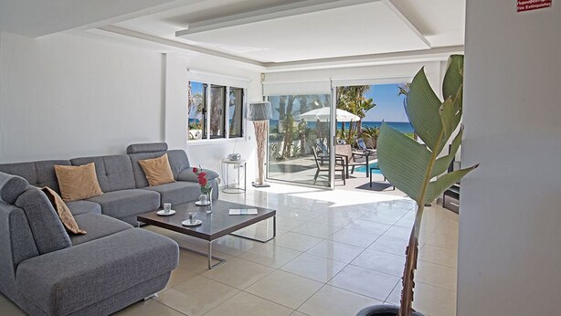 Una sala de estar moderna con un sofá gris, una mesa de centro y un ventanal con vistas a una piscina y al mar.