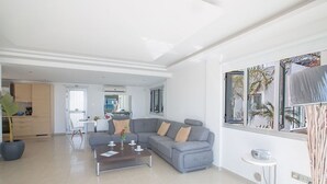 Amplia sala de estar con un sofá seccional gris, una mesa de centro y vistas a palmeras a través de grandes ventanales.