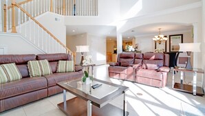 A spacious living area with a brown leather sofa, a glass coffee table, and a pink leather sofa. A wooden staircase with white balusters leads to an upper level. - Imagine You and Your Family Renting This 5 Star Villa on Providence Resort, Orlando Villa 3629 (Davenport)