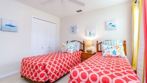 Une chambre avec deux lits simples, chacun recouvert d’une couvre-lit rouge et blanche à motifs circulaires, doté d’une tête de lit en bois et agrémentée d’une table de chevet avec une lampe. Les murs sont décorés d’œuvres d’art représentant la vie marine, et une porte blanche ferme la pièce.