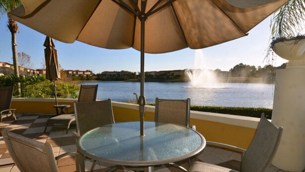 A patio with a round glass table and chairs, an umbrella, and a view of a lake with a fountain. - Perfect Townhome on Enchantada Resort Orlando Townhome 5495 (Kissimmee)