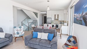 A modern living room with a grey sofa, a colorful armchair, and a dining area with a kitchen in the background. - Luxury 5 Bedroom Villa With Private Pool, Protaras Villa 1250 (Paralimni)
