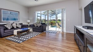 A spacious living room with a dark-colored sofa, a coffee table, and a flat-screen TV. There is a large window offering a view of a swimming pool and greenery. - Luxury 5 Bed Villa, Perfect for Large Groups, 1005 (Davenport)