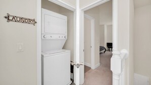 Une buanderie équipée d’un lave-linge et d’un sèche-linge, une porte avec une pancarte indiquant « buanderie », et un couloir menant à une chambre à coucher avec un lit et une chaise.