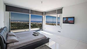 Ein modernes Schlafzimmer mit einem großen Bett, einem an der Wand montierten Fernseher und Blick auf die Stadt durch große Fenster.