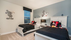 Une chambre avec deux lits, une fenêtre avec des stores, une table de chevet, une lampe et une décoration sur le thème de Mickey Mouse.