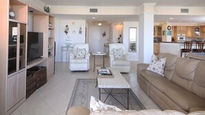 A spacious living room with a beige sofa, two armchairs, a coffee table, and a TV. There is a kitchen in the background with wooden cabinets and a clock. - Perfect Apartment on The Anchorage, Siesta Key Apartment 1022 (Siesta Key)