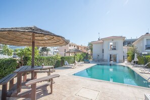 Área de piscina com guarda-sol de palha, bancos de madeira e um edifício de dois andares ao fundo.