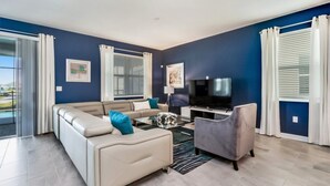 A modern living room with a beige sofa, a grey armchair, a glass coffee table, and a flat-screen TV. There are two windows with white curtains and a view of the outdoors. - Rent a Luxury Mansion on Storey Lake Resort, Minutes From Disney, Orlando Mansion 3232 (Kissimmee)
