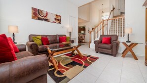 A spacious living room with a brown leather sofa, a glass coffee table, and a patterned rug. A wooden chair and a small table with a lamp are also visible.