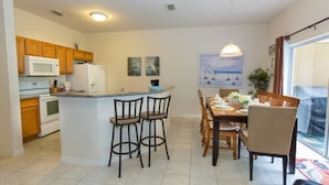 A compact kitchen with a central island, wooden cabinets, a microwave, and a refrigerator. There are two bar stools and a dining table with chairs. - Rent a Luxury Villa on Encantada Resort, Minutes From Disney, Orlando Villa 1062 (Kissimmee)