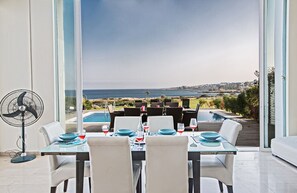 Uma área de jantar com mesa posta para quatro, com vista para uma piscina e o mar.