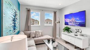 Ein modernes Wohnzimmer mit einem beigefarbenen Sofa, einem Glastisch und einem weißen Fernseher an der Wand, der 'Walt Disney Pictures' zeigt.