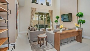 Ein geräumiges Wohnzimmer mit einer Couch, einem Couchtisch und einem Fernseher. Pflanzen und ein Fenster mit Vorhängen sind ebenfalls vorhanden.