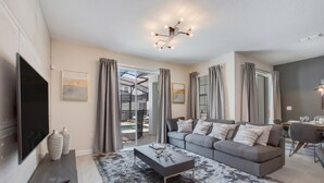 A modern living room with a grey sofa, a coffee table, and a TV. There are curtains, a chandelier, and a dining area visible. - Rent a Luxury Townhome on Storey Lake Resort, Minutes From Disney, Orlando Townhome 3093 (Kissimmee)