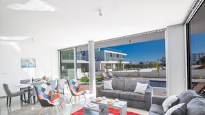 A modern living room with a pool view, grey sofas, a red rug, and a dining area with chairs and a table. - Family Bliss Awaits: Protaras Villa 1572 (Paralimni)