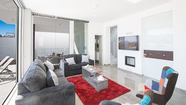 A modern living room with a grey sofa, a red rug, and a dining area with a table and chairs. - Family Bliss Awaits: Protaras Villa 1572 (Paralimni)
