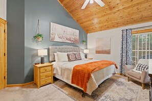 Quarto com cama, mesas de cabeceira, criadeira, uma cadeira e janela com cortinas.