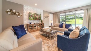 A modern living room with a beige sofa, a blue armchair, and a beige couch. There is a coffee table in the center, a TV on the wall, and a large window with a view of a garden. - Rent the Perfect 8 Bedroom Villa on Encore Resort at Reunion, Orlando Villa 4474 (Kissimmee)