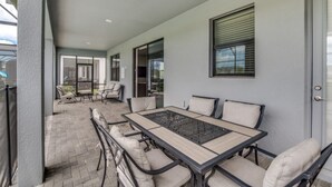 Ein überdachter Patio-Bereich mit einem langen Tisch und Stühlen, eine Glastür, die zu einem Innenbereich führt, sowie ein Fenster mit Jalousien.