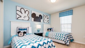 Un dormitorio con dos camas, cada una con ropa de cama temática de Mickey Mouse. Hay cuadros en las paredes sobre las camas y una ventana con persianas.