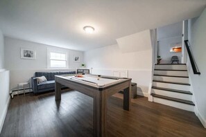 A modern living room with a pool table, a grey sofa, a staircase, and a small table with a plant.