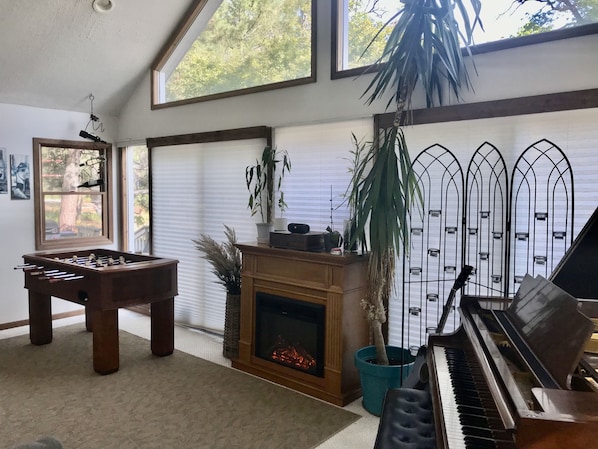 A cozy living room with a foosball table, a fireplace, a piano, and large windows.