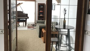 A room with a piano, a dining table with chairs, a bookshelf, and a small table with a lamp.