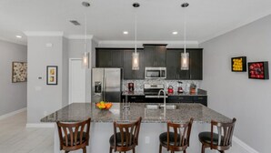 A modern kitchen with granite countertops, stainless steel appliances, and dark wooden cabinets. There are four wooden chairs around the island, and pendant lights hang above.